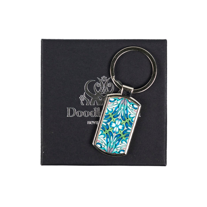 A metal keyring with a millefiori floral pattern inlay, attached to a black keychain fob that reads &